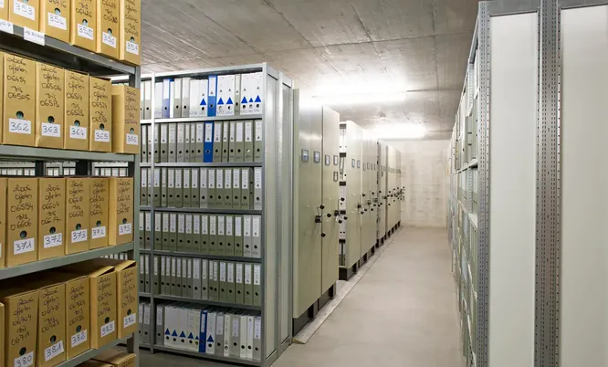 Aktenlager mit zahlreichen Archivkartons, Ordnern und verschiebbaren Regalsystemen zur langfristigen Aufbewahrung von Dokumenten.