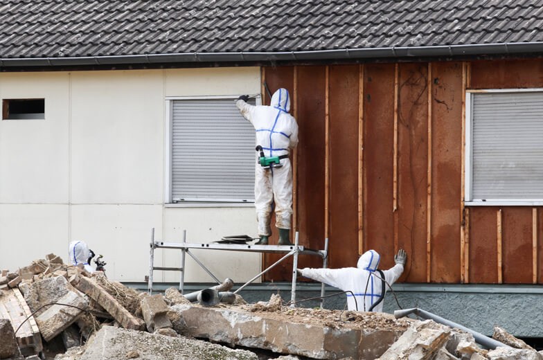 Fachpersonal in Schutzanzügen führt Sanierungsarbeiten an einer Hausfassade durch, Entfernung schadstoffbelasteter Materialien im Außenbereich.
